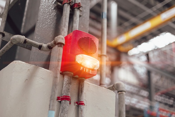 A red fire alarm with a flashing light is installed on a wall inside an industrial building.
