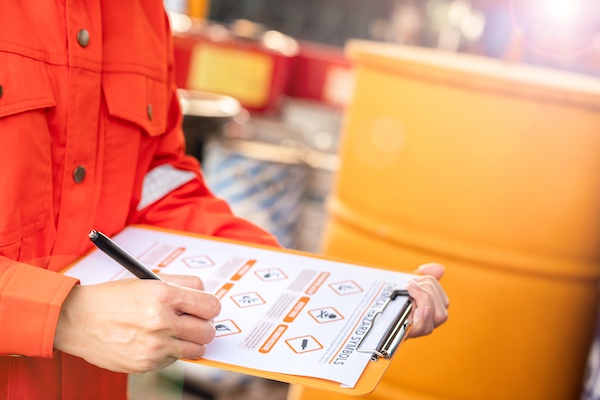 A person in orange safety gear writes on a clipboard beside yellow chemical barrels marked with hazard symbols.