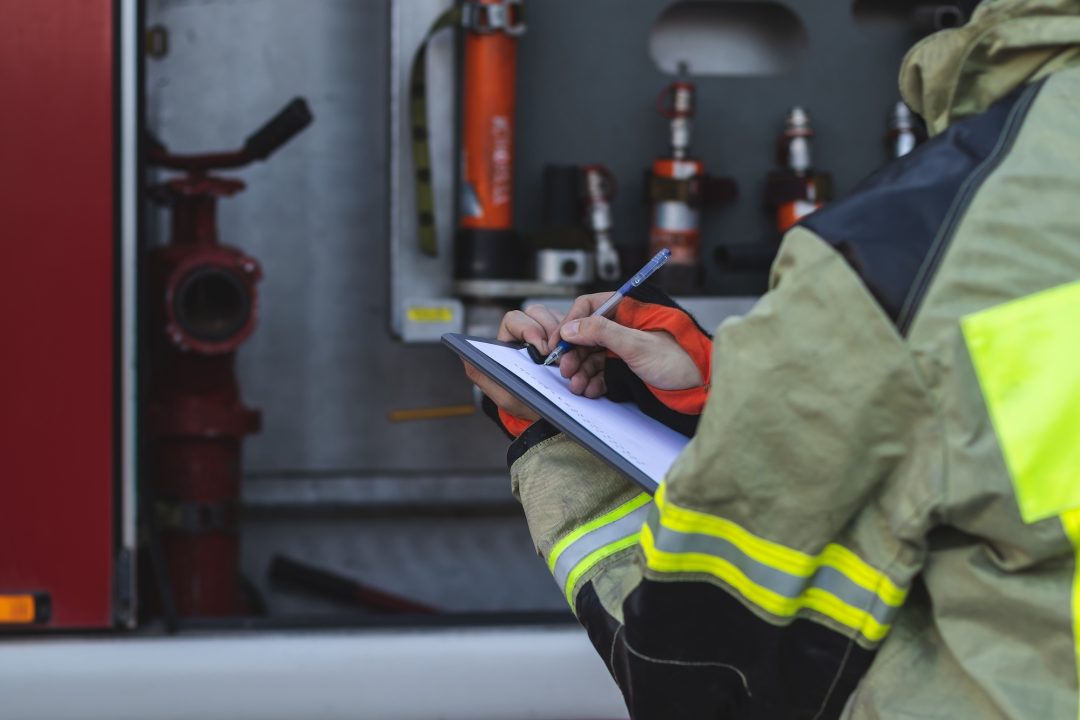 A firefighter wearing full gear writes on a clipboard with various fire equipment and tools visible in the background.